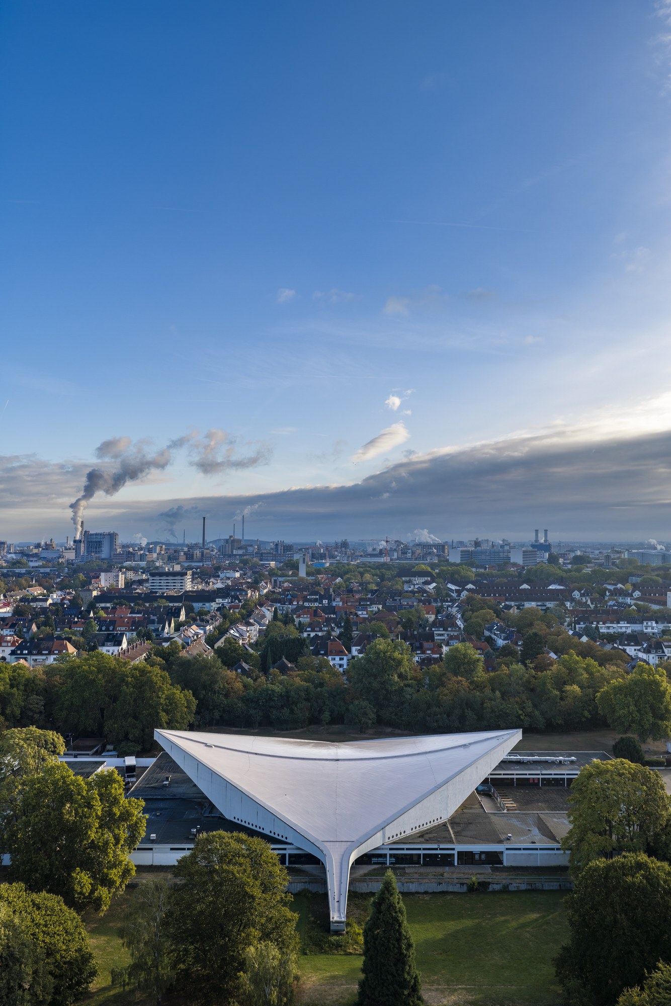 Friedrich-Ebert-Halle, Ludwigshafen am Rhein — das geschwungene Dach im Grün des Ebertparks, dahinter Stadt und Kraftwerksschlote