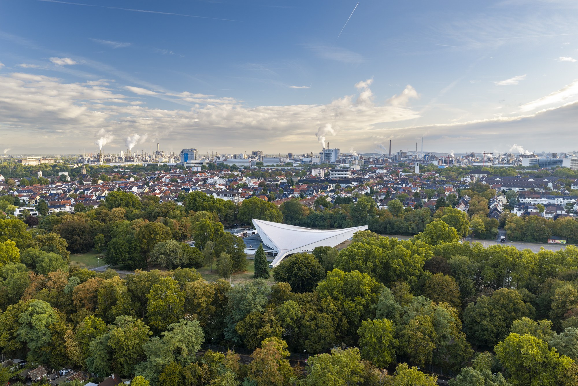 Friedrich-Ebert-Halle, Ludwigshafen am Rhein — die Hyparschale über den Baumkronen des Ebertparks, dahinter die Industriesilhouette der BASF