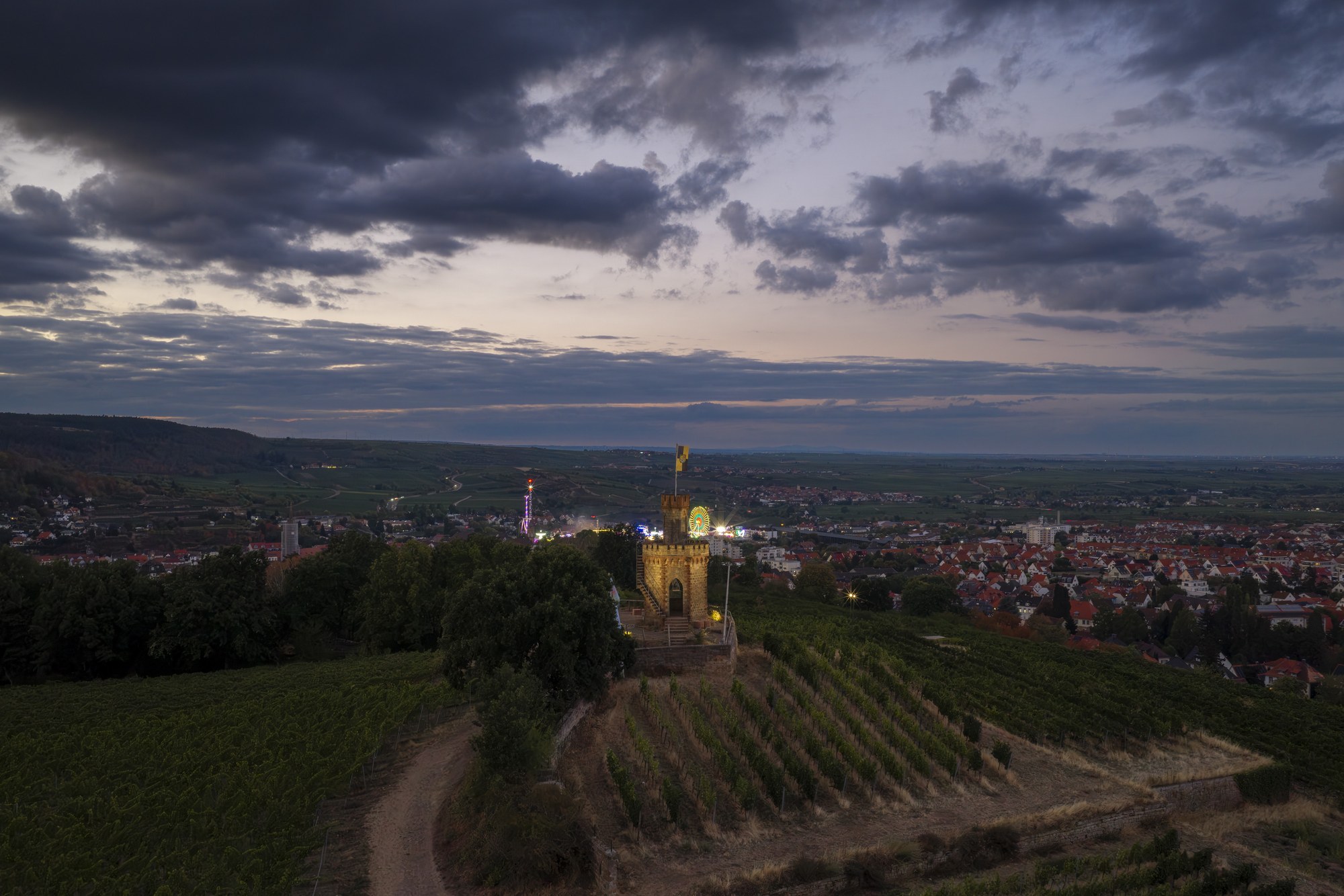 Flaggenturm auf dem Fuchsmantel, Bad Dürkheim — Luftbild über die Weinberge zum beleuchteten Turm, im Hintergrund die Lichter des Wurstmarkts