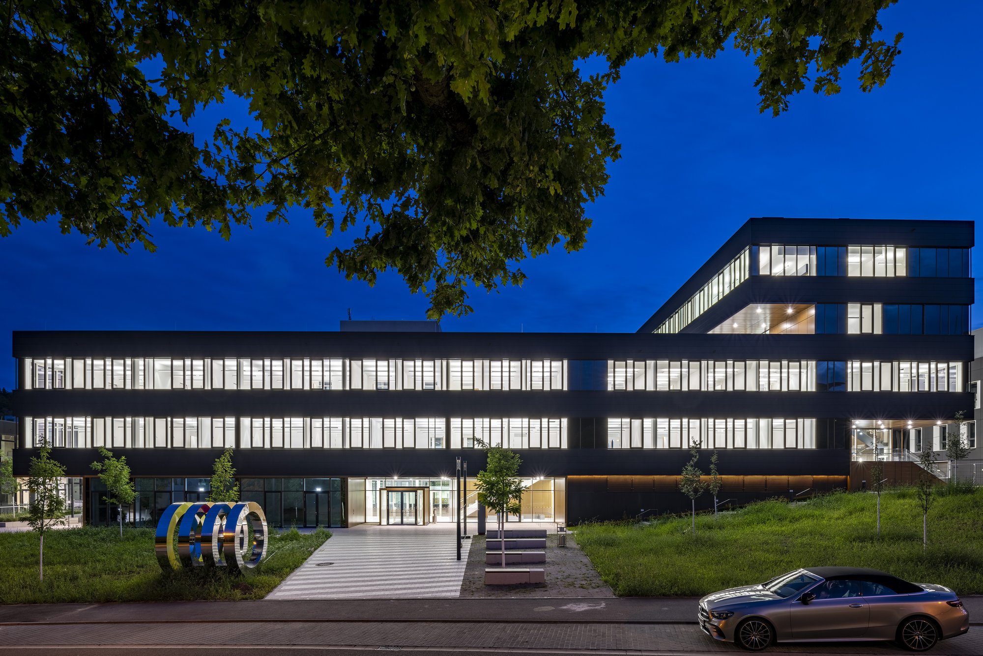 Architekturfotografie Zentrum für Biophysik ZBP, Saarbrücken — Blaue Stunde, Blick von der Straße über den Vorplatz zum beleuchteten Eingang