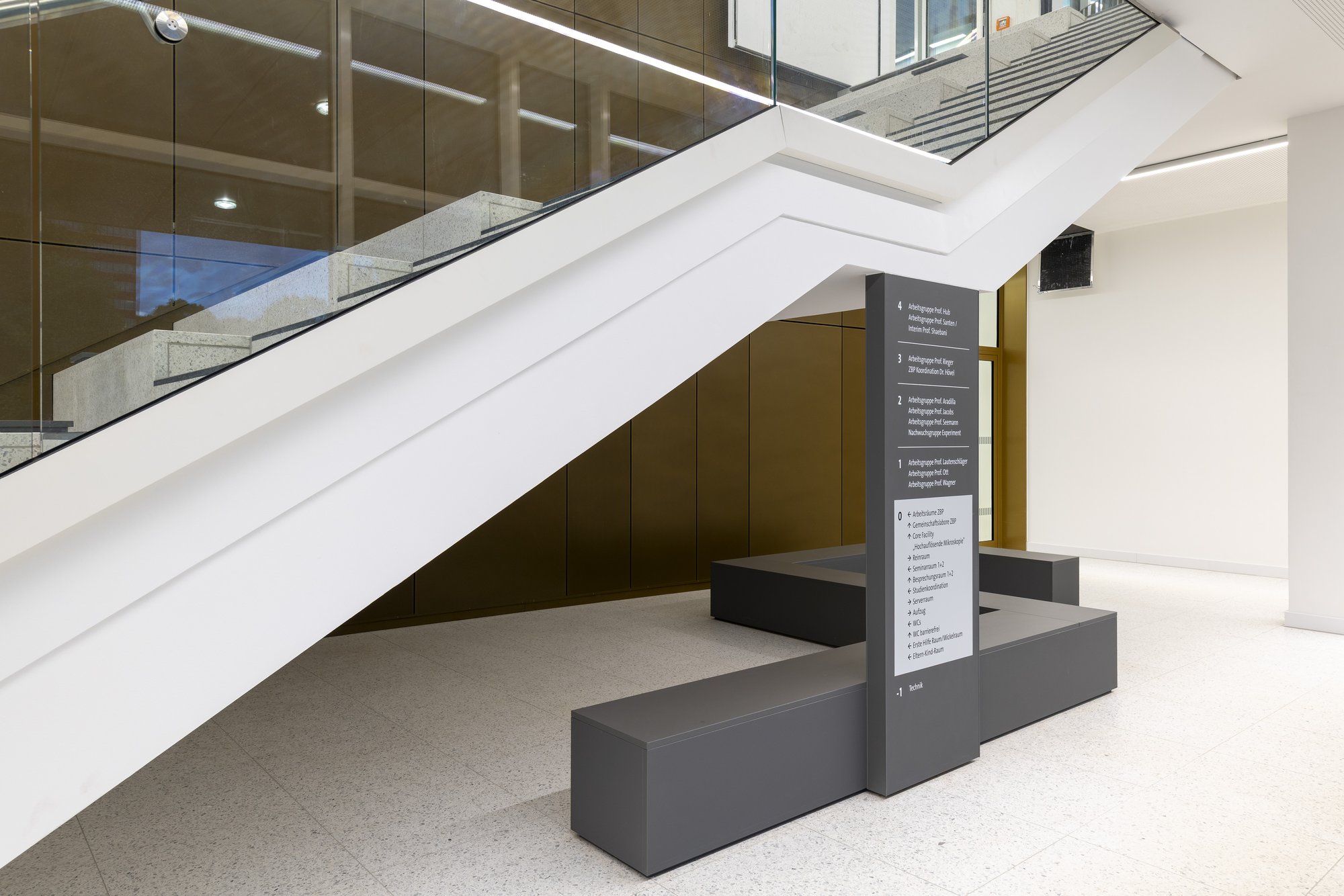 Architekturfotografie Zentrum für Biophysik ZBP, Saarbrücken — Foyer mit der weiß verkleideten Treppenuntersicht, Leitsystemstele und Sitzbänken