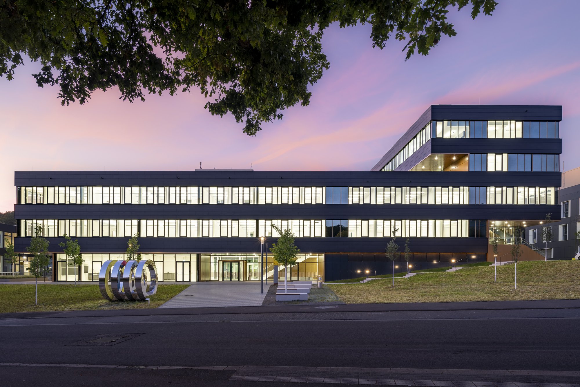 Architekturfotografie Zentrum für Biophysik ZBP, Saarbrücken — Der beleuchtete Neubau am Abend, gerahmt von den Ästen eines Baums