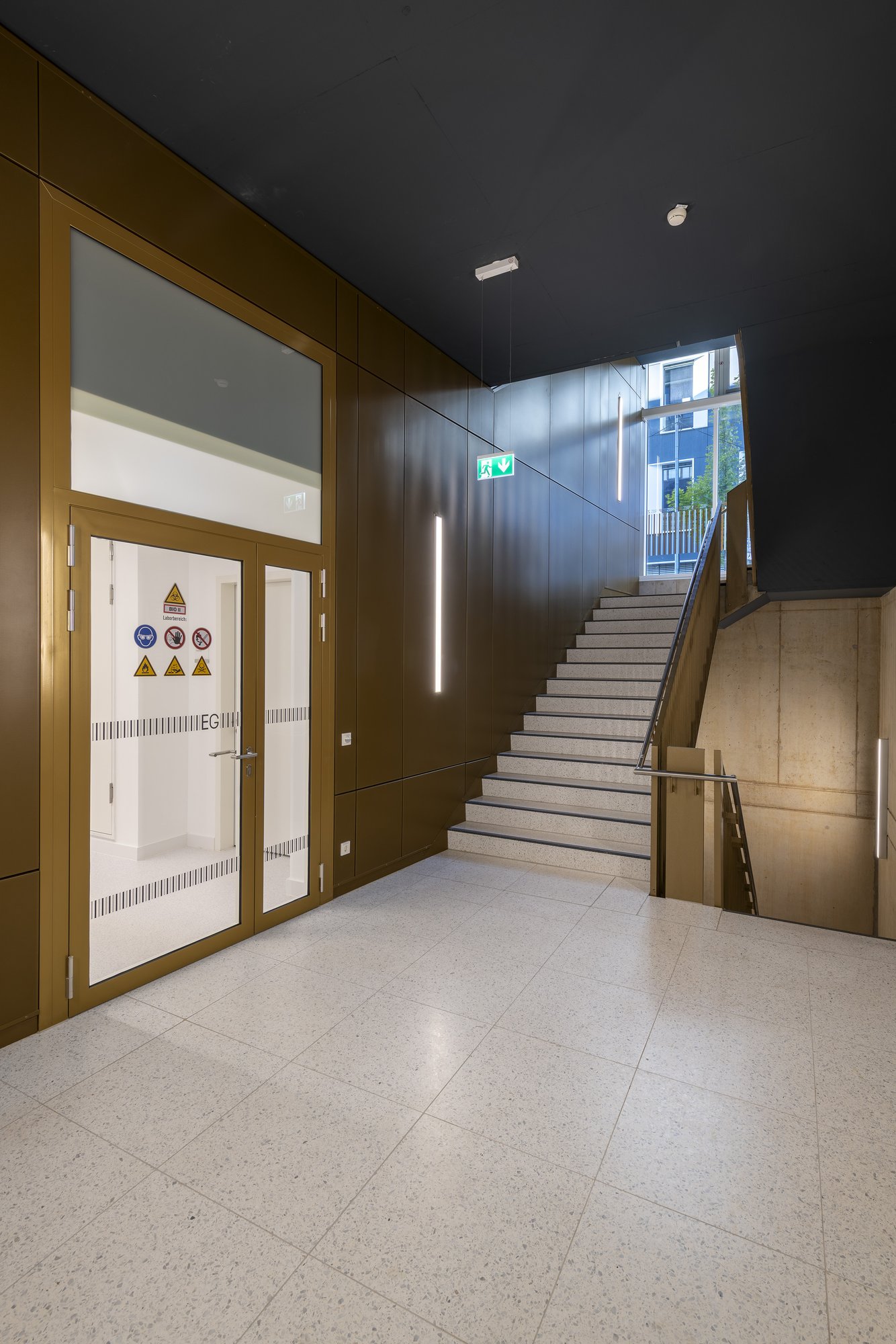 Architekturfotografie Zentrum für Biophysik ZBP, Saarbrücken — Treppenhaus mit dunkler Decke, bronzefarbener Wandbekleidung und verglaster Labortür