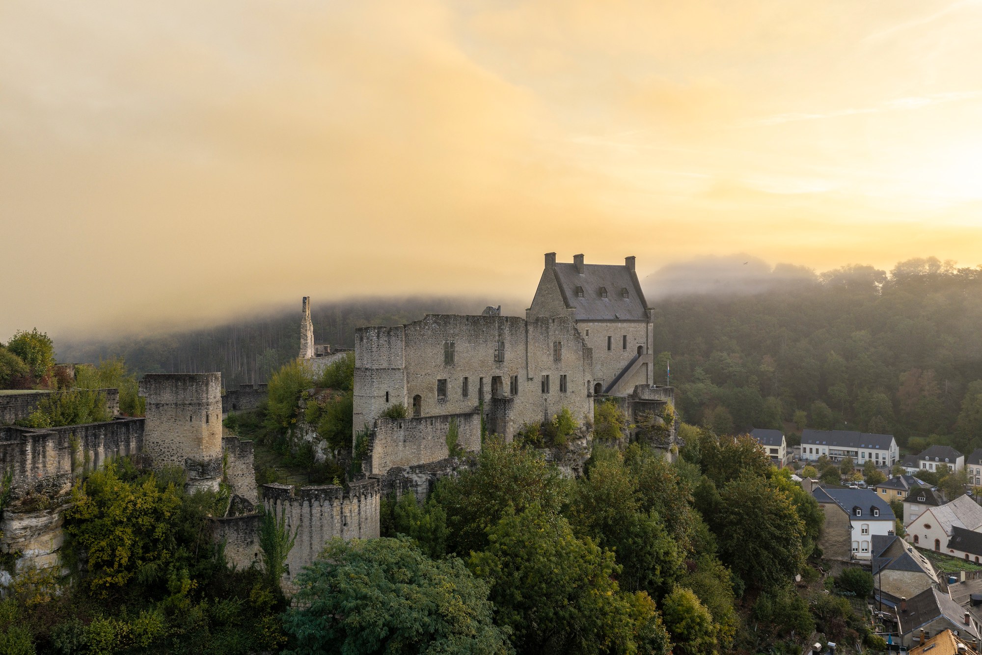 Architekturfotografie Burg Larochette — Luftbild der Ruinen im Gegenlicht der aufgehenden Sonne, Nebel über dem Tal, rechts unten der Ort