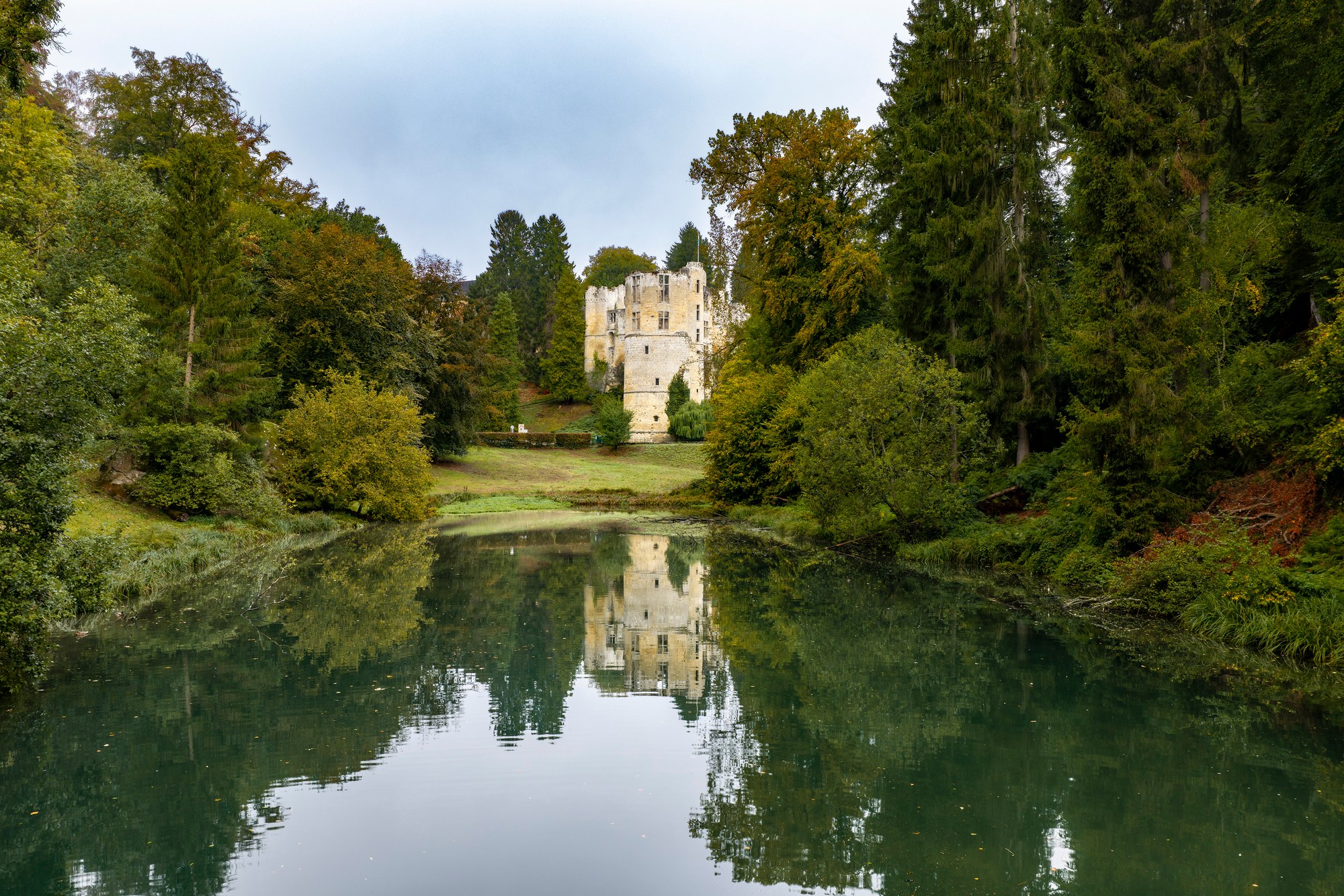 Architekturfotografie Burg Beaufort — die Ruine am Ende des Weihers, gespiegelt im Wasser, gerahmt von Bäumen