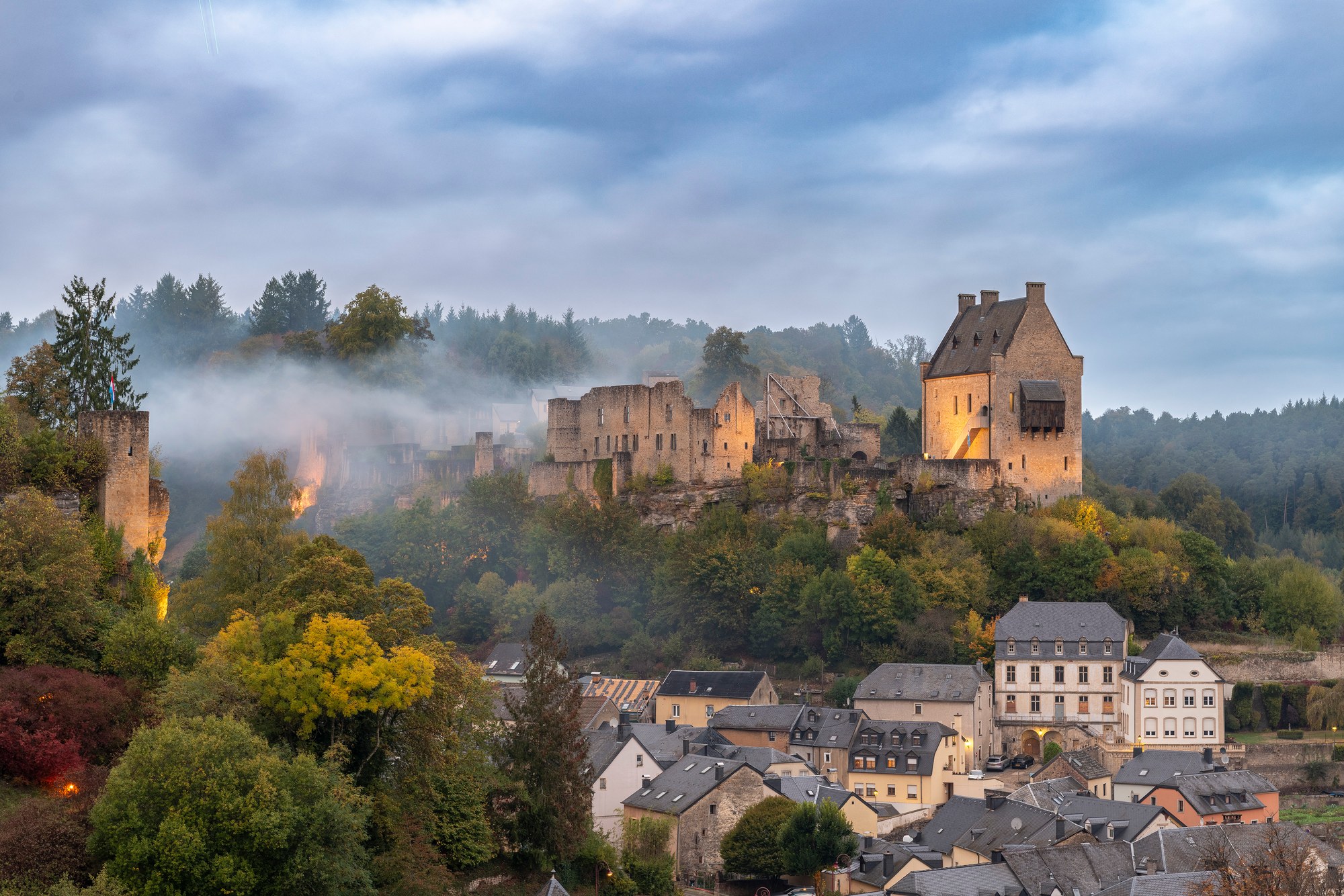 Architekturfotografie Burg Larochette — die Burg über den Dächern des Orts in der Abenddämmerung, das Criechinger Haus beleuchtet, Nebel über dem Wald