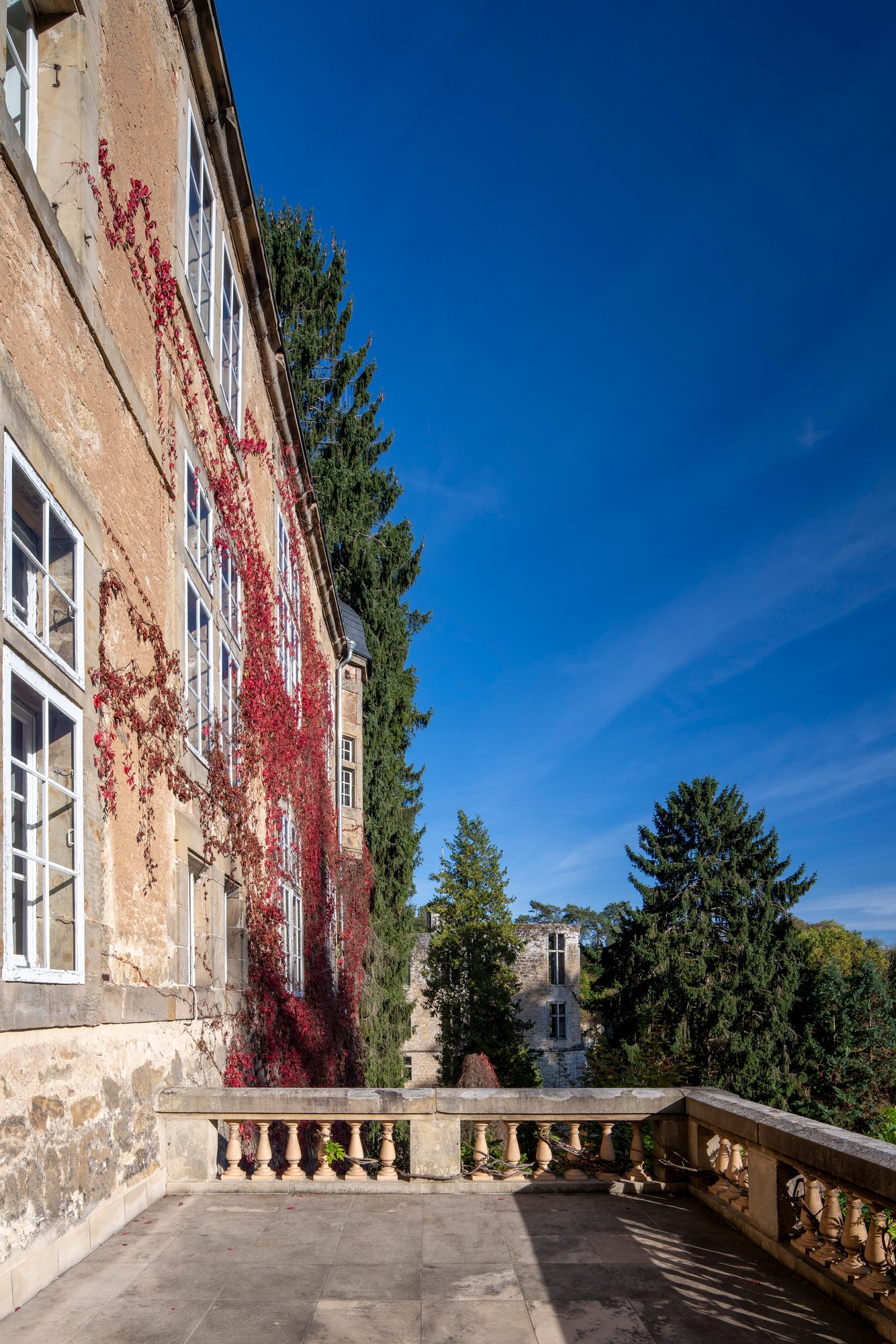 Architekturfotografie Renaissanceschloss Beaufort — Terrasse mit Balustrade vor der Fassade mit rotem Wildem Wein, im Hintergrund die Burg